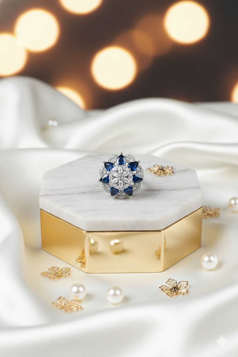 Gold ring with blue gemstones on a white marble box with pearls around, blurred lights in the background